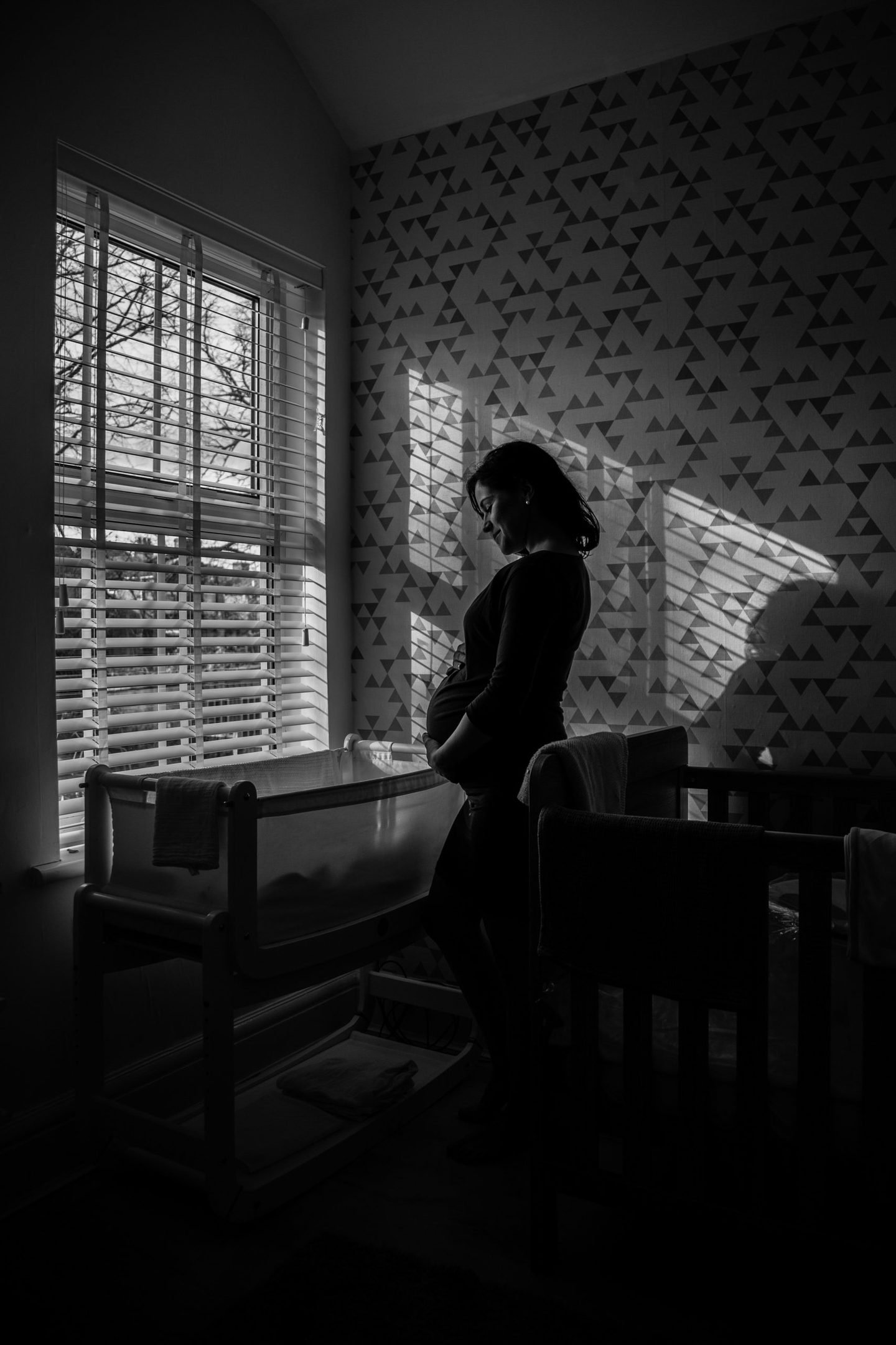 Black and white silhouette of an expectant mother standing by a nursery window, light falling through the blinds