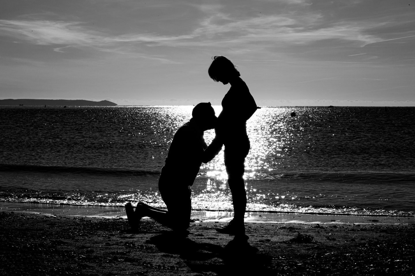 Silhouette on a beach at sunset of a kneeling father kissing his partner's pregnant bump
