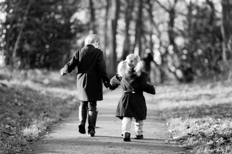Black and white photograph of two young siblings in coats holding hands as they walk down a woodland path