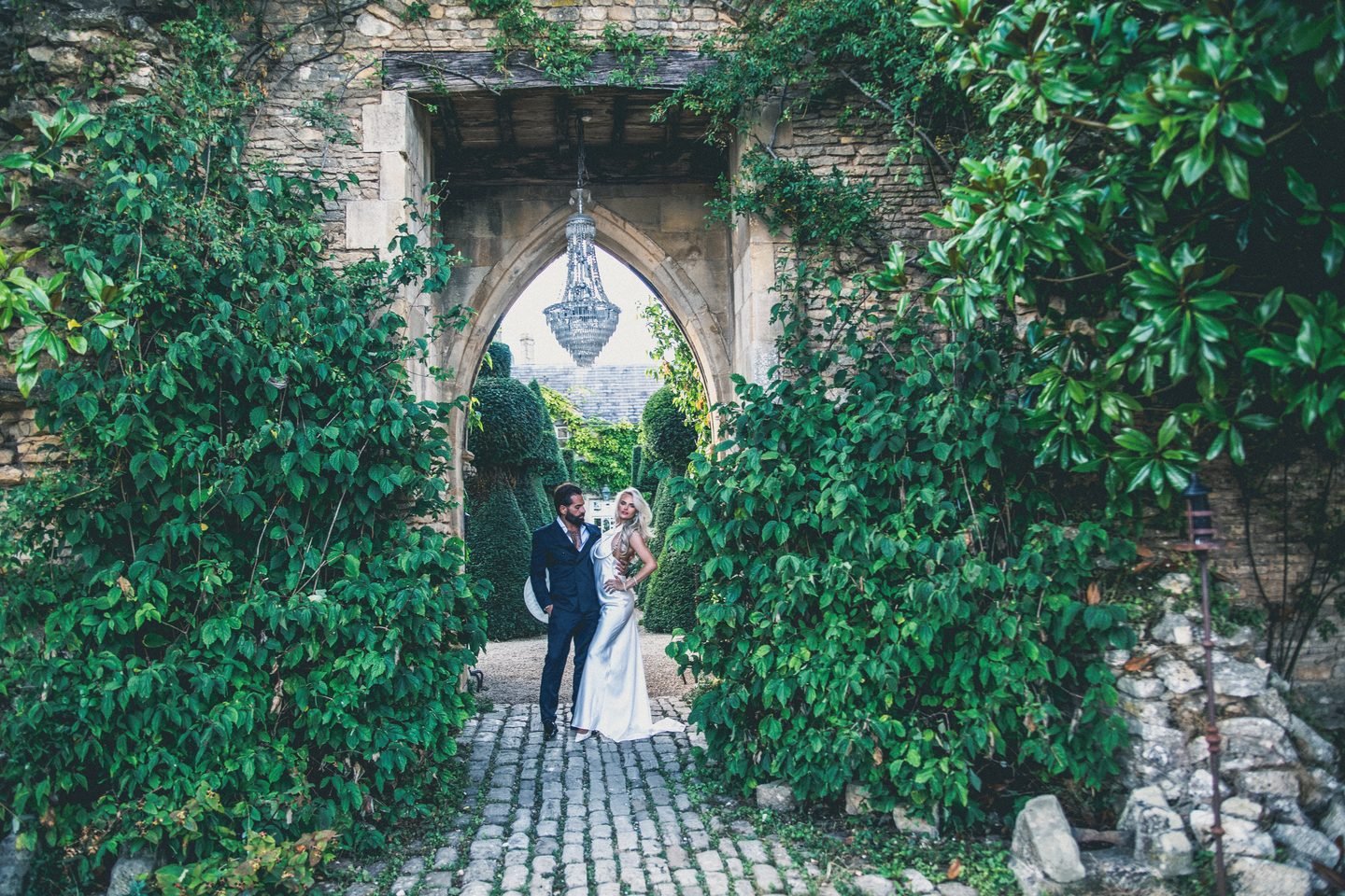 Bride and groom framed by an ivy-covered gothic stone archway with a chandelier hanging above a cobbled path