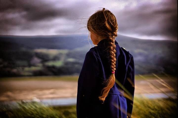 A young girl with a long plaited braid gazes across moody hills and countryside