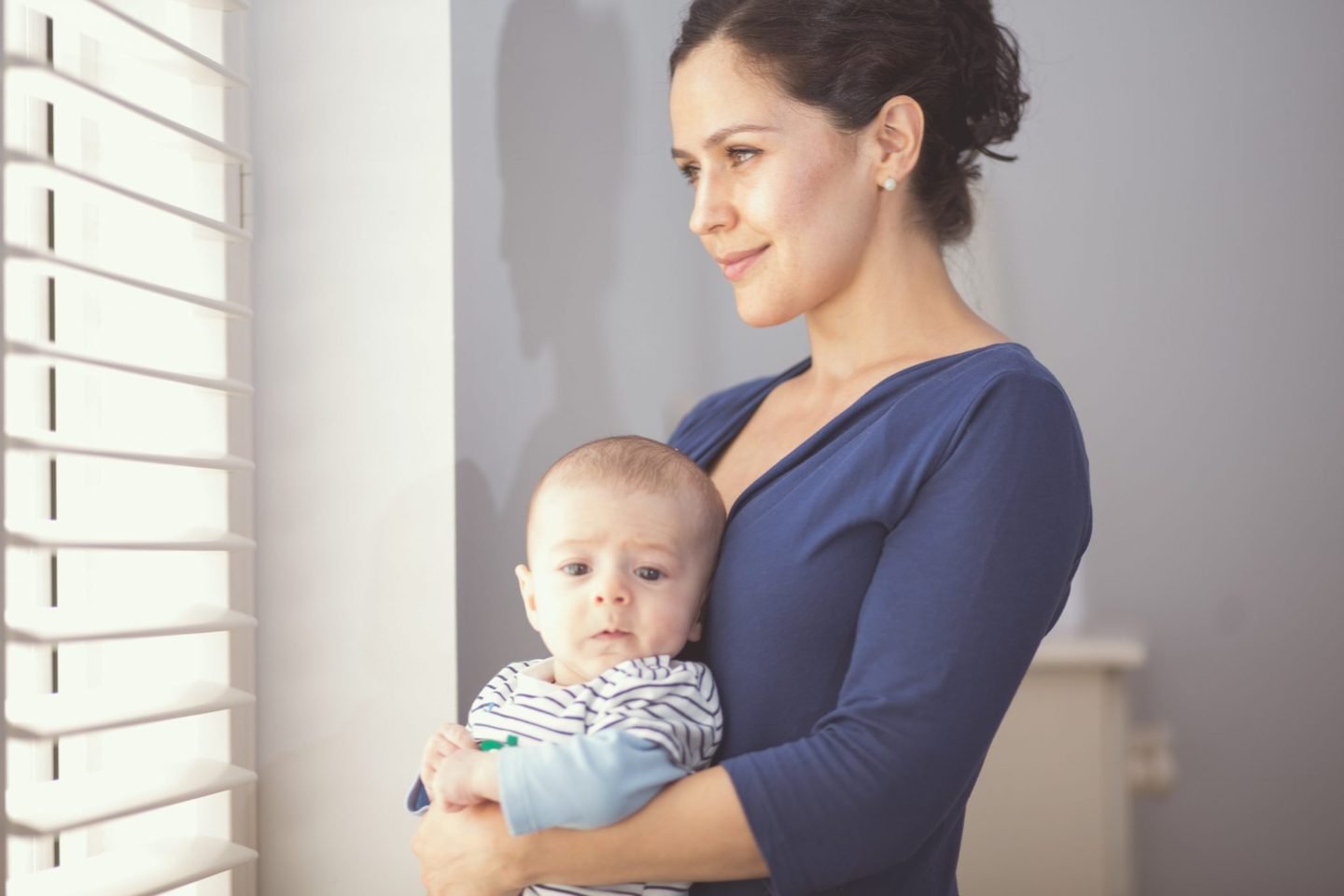 A mother in a blue dress holds her baby son beside a sunlit window