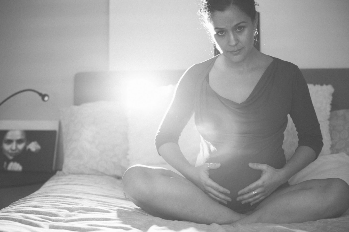 Black and white maternity portrait of a woman sitting cross-legged on a bed, hands on her bump, backlit by a lamp