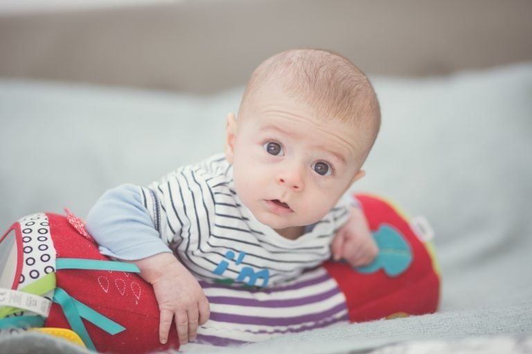 A baby in a striped top lifts his head during tummy time on a colourful cushion