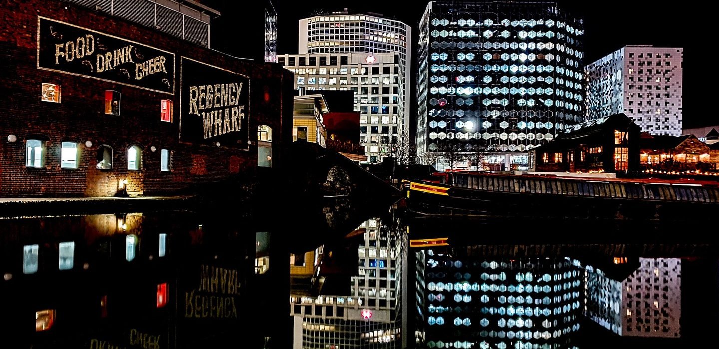 Regency Wharf in Birmingham at night, its signage and city towers reflected in the canal