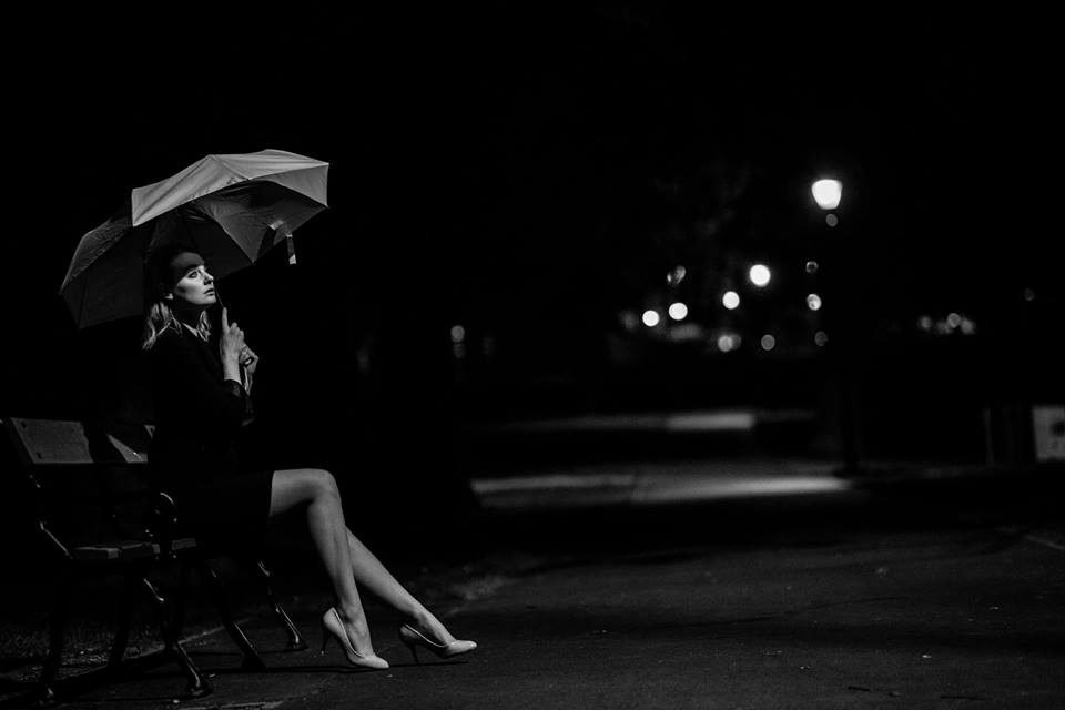 Black and white night portrait of a woman with an umbrella seated on a park bench under street lamps