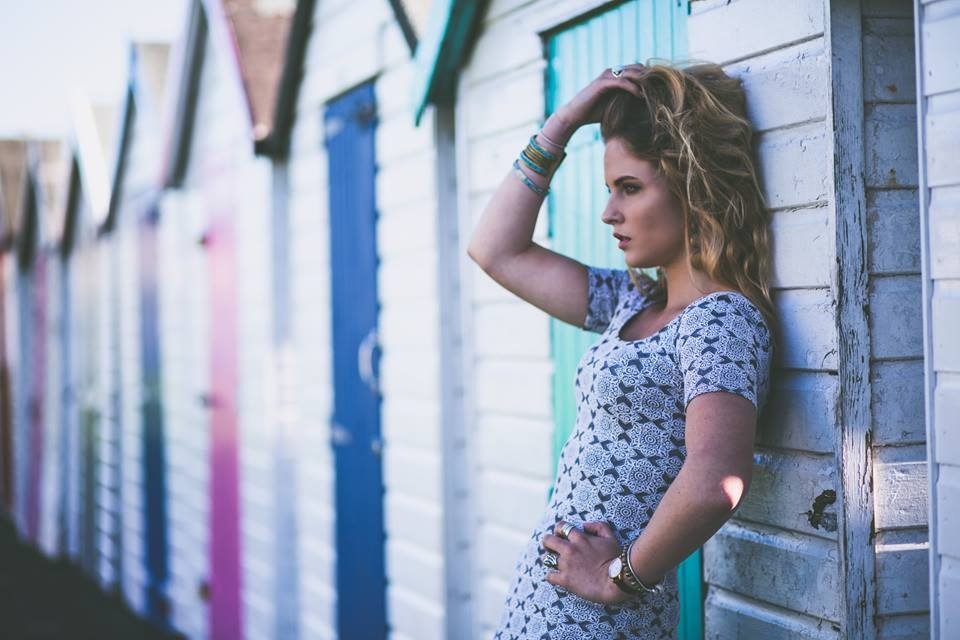 A woman in a patterned dress leans against colourful beach huts in natural light