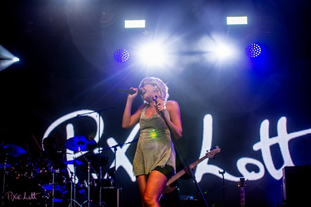 Pixie Lott singing on a festival stage under blue lights in front of her illuminated name