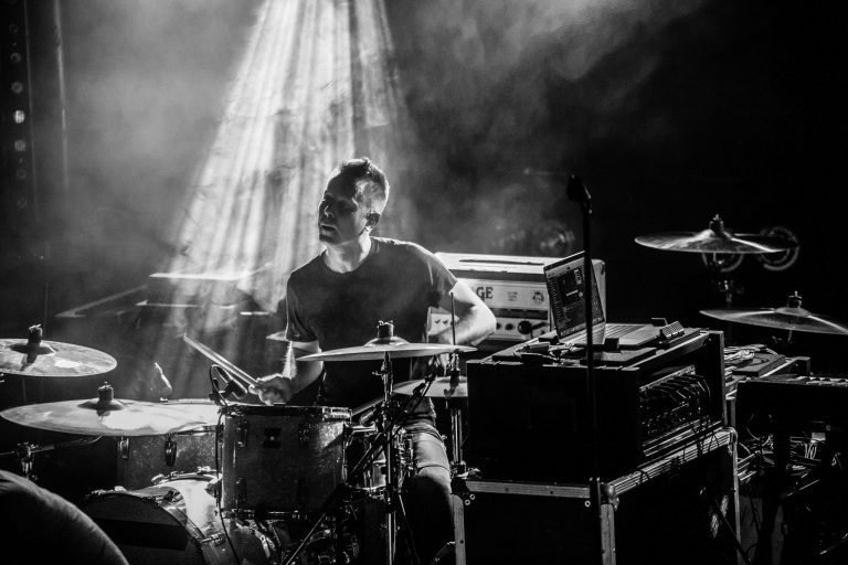 Black and white photograph of a Mogwai drummer behind the kit in shafts of stage light and haze