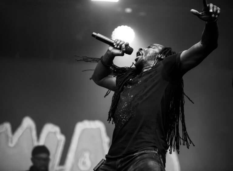 Black and white photograph of a singer with long dreadlocks, arm raised, singing into the lights