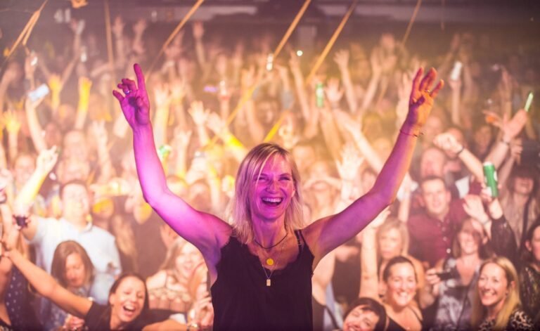 Jo Whiley with arms raised in front of a packed crowd with hands in the air at a 90s anthems night in Birmingham