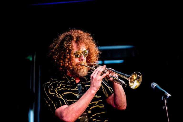 A curly-haired trumpet player in sunglasses performing on stage