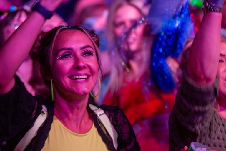 A smiling woman with glitter make-up dancing in the crowd under pink light at Ibiza Proms, Solihull