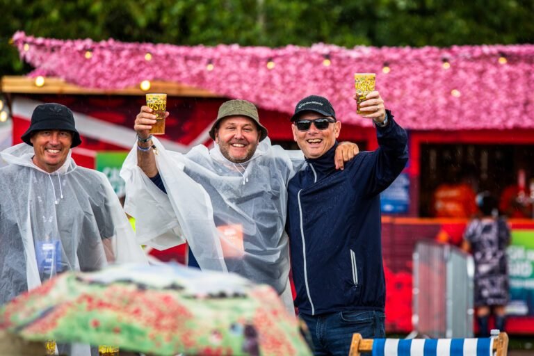 Three men in rain ponchos raising pints in front of a flower-covered bar at Solihull Summerfest