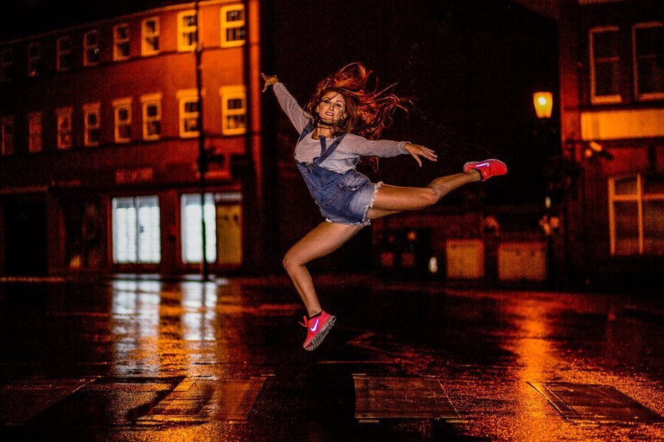 A dancer leaping on a wet street at night, lit orange by street lights