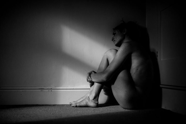 Fine art black and white portrait of a woman sitting against a wall in window light and shadow
