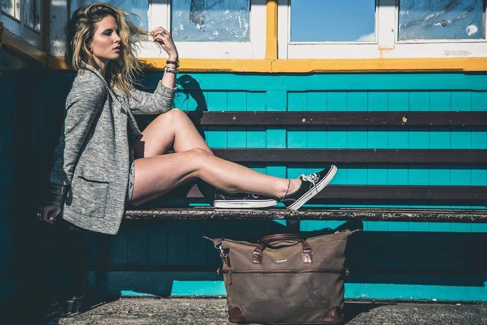 A woman in a grey blazer sitting on a turquoise seaside bench with a leather holdall