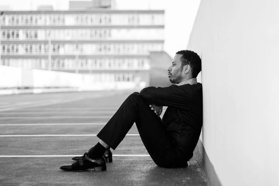 Black and white portrait of a man in a suit sitting against a wall on a car park roof