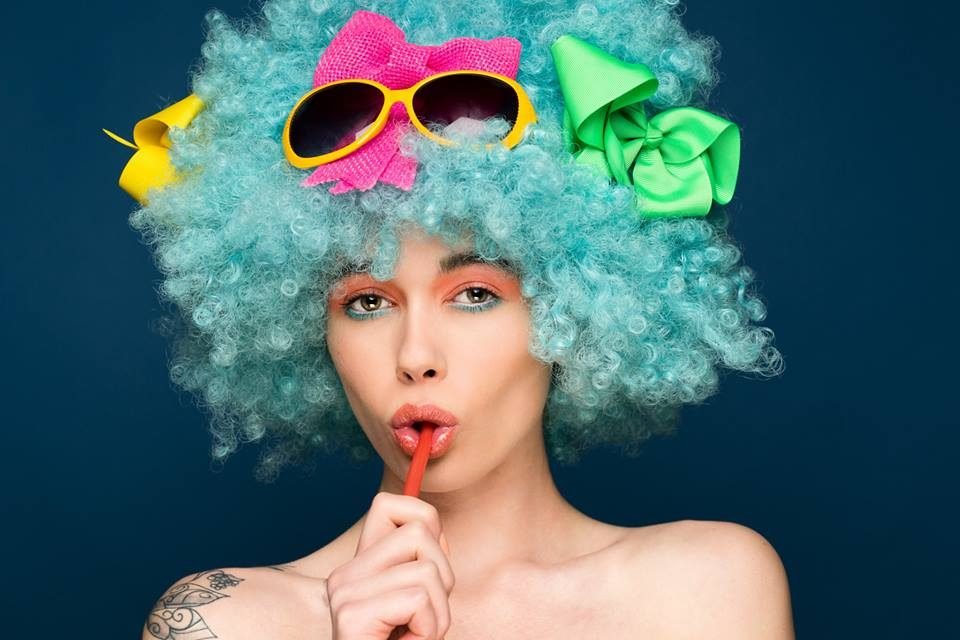 Colourful pop art studio portrait of a woman in a blue curly wig, bows and sunglasses with a red lollipop