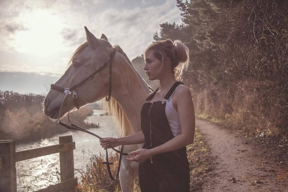 A woman in dungarees standing with a white horse beside a river path in golden light