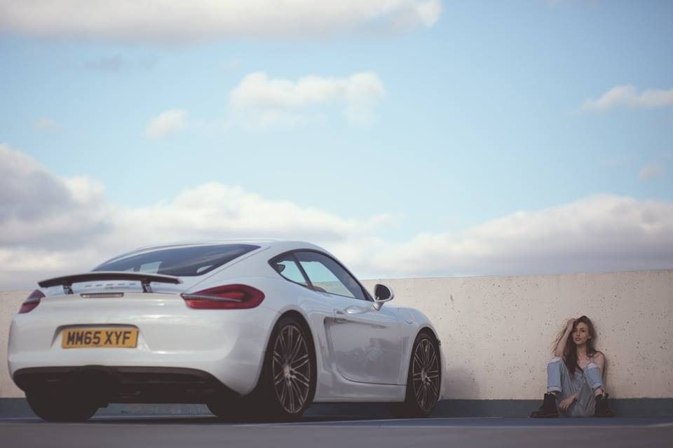 A woman sitting against a rooftop wall beside a white sports car under a blue sky