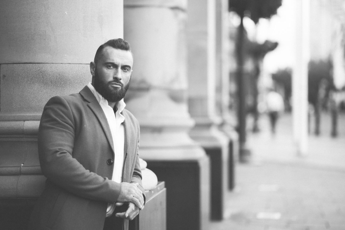 Black and white portrait of a bearded man in an overcoat leaning against stone columns