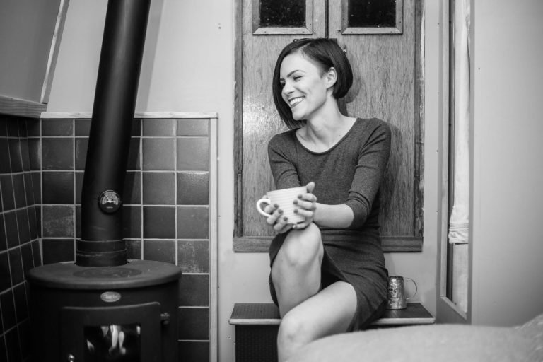 Black and white candid portrait of a smiling woman with a mug beside a wood burner