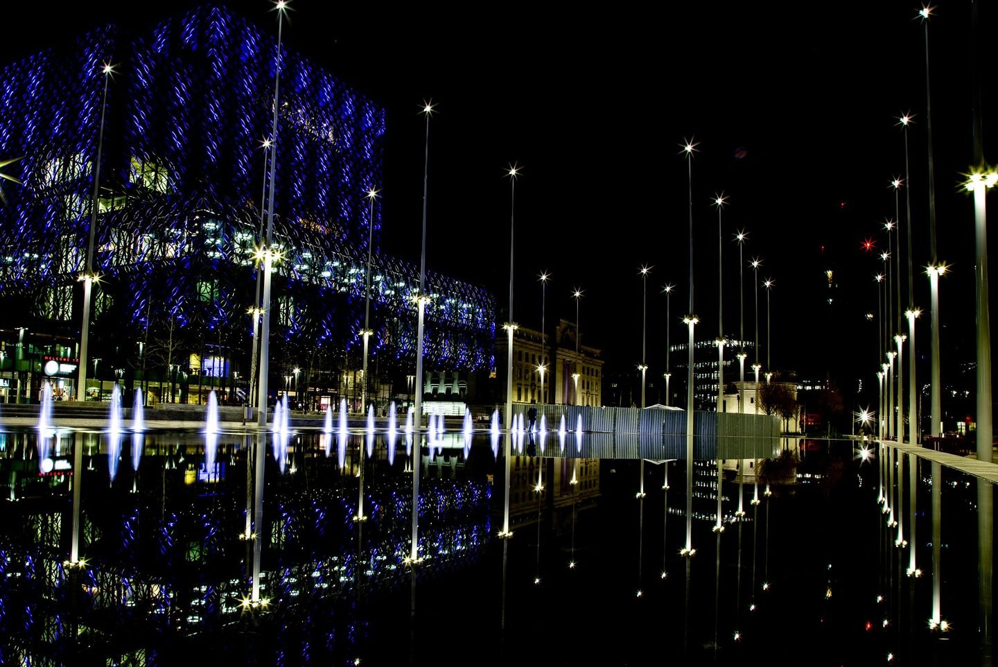 The Library of Birmingham's blue lit facade reflected in the water features of Centenary Square at night
