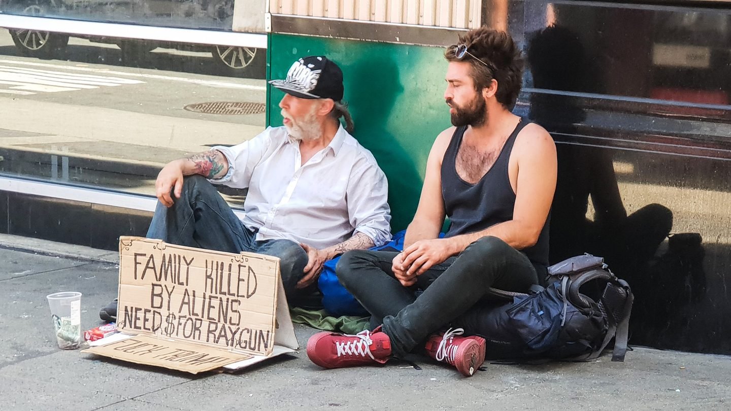 Two men sitting on a Manhattan pavement beside a humorous cardboard sign