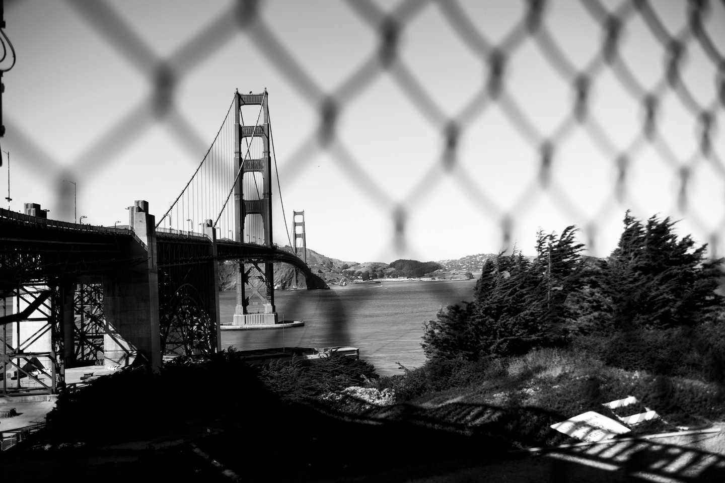 Black and white photograph of the Golden Gate Bridge seen through chain link fence