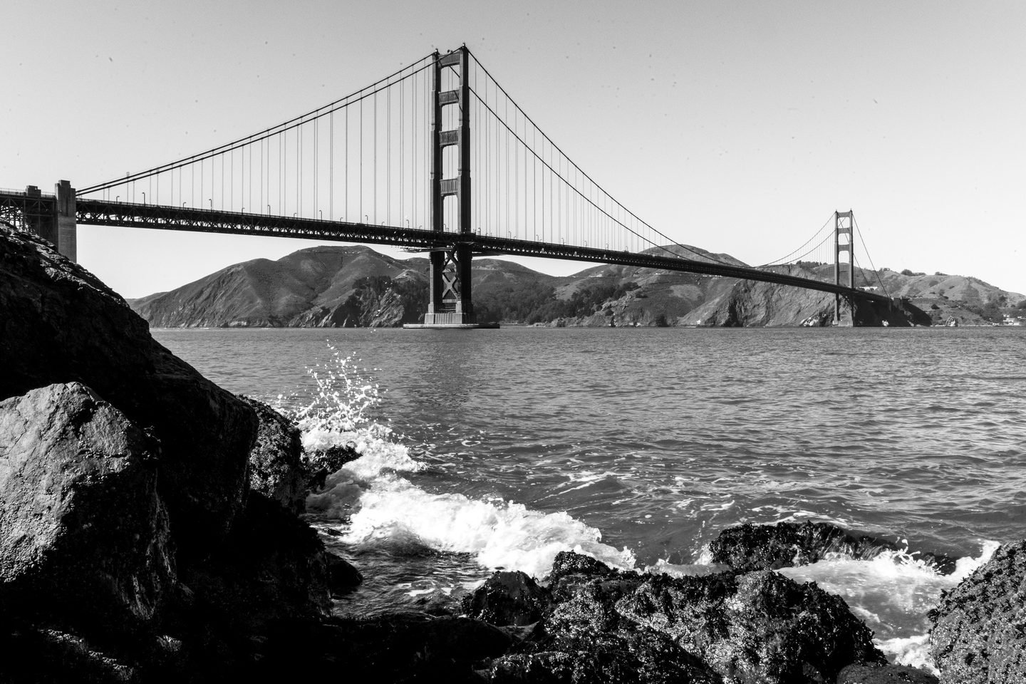 Black and white photograph of waves breaking on rocks below the Golden Gate Bridge